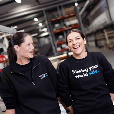 Two smiling Troax employees in branded fleece jackets with the slogan 'Making your world safe.' sharing a light moment in an industrial setting.