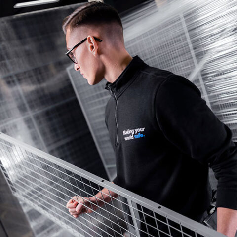 A professional wearing a black Troax fleece with the slogan 'Making your world safe.' stands beside a safety mesh partition, symbolizing commitment to secure environments.